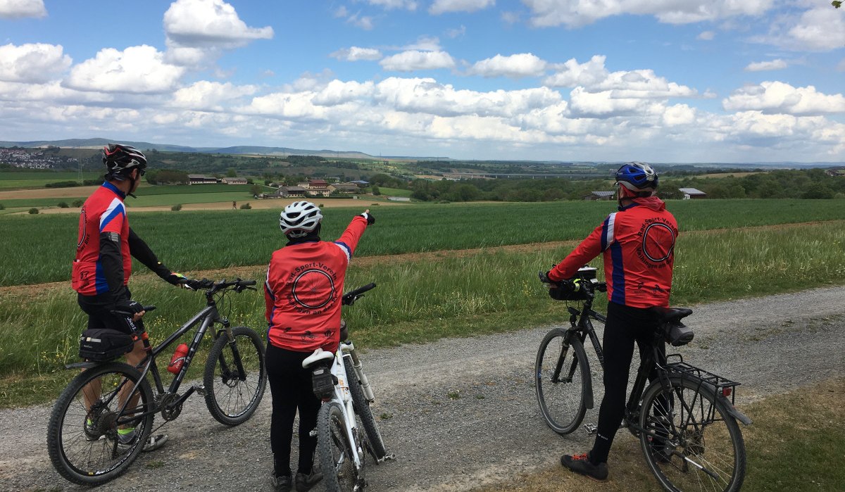 Drei Radfahrer in roten Trikots stehen auf einem Feldweg und schauen in die weite Landschaft. Einer zeigt in die Ferne. Der Himmel ist bewölkt., © Land der 1000 Hügel - Kraichgau-Stromberg Drei Radfahrer in roten Trikots stehen auf einem Feldweg und schauen in die weite Landschaft. Einer zeigt in die Ferne. Der Himmel ist bewölkt., © Land der 1000 Hügel - Kraichgau-Stromberg