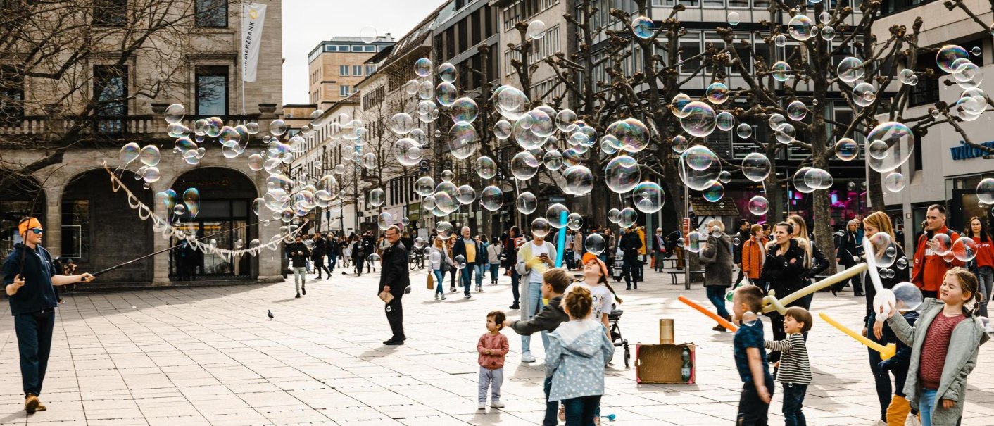Ein Straßenkünstler erzeugt Seifenblasen auf einer belebten Straße. Kinder spielen begeistert mit den Blasen, während Passanten zuschauen., © Stuttgart-Marketing GmbH, Sarah Schmid