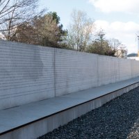 Eine lange Betonwand mit eingravierten Namen steht neben stillgelegten Bahngleisen unter einem bewölkten Himmel., © Foto-Kunst Andreas Keller Eine lange Betonwand mit eingravierten Namen steht neben stillgelegten Bahngleisen unter einem bewölkten Himmel., © Foto-Kunst Andreas Keller