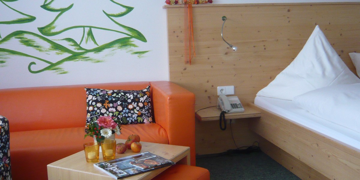Hotelzimmer mit orangefarbenem Sofa, Holzbett, Blumen auf Tisch, Zeitschrift, Telefon und gr&uuml;ner Wandmalerei., &copy; Hotel Hochwald