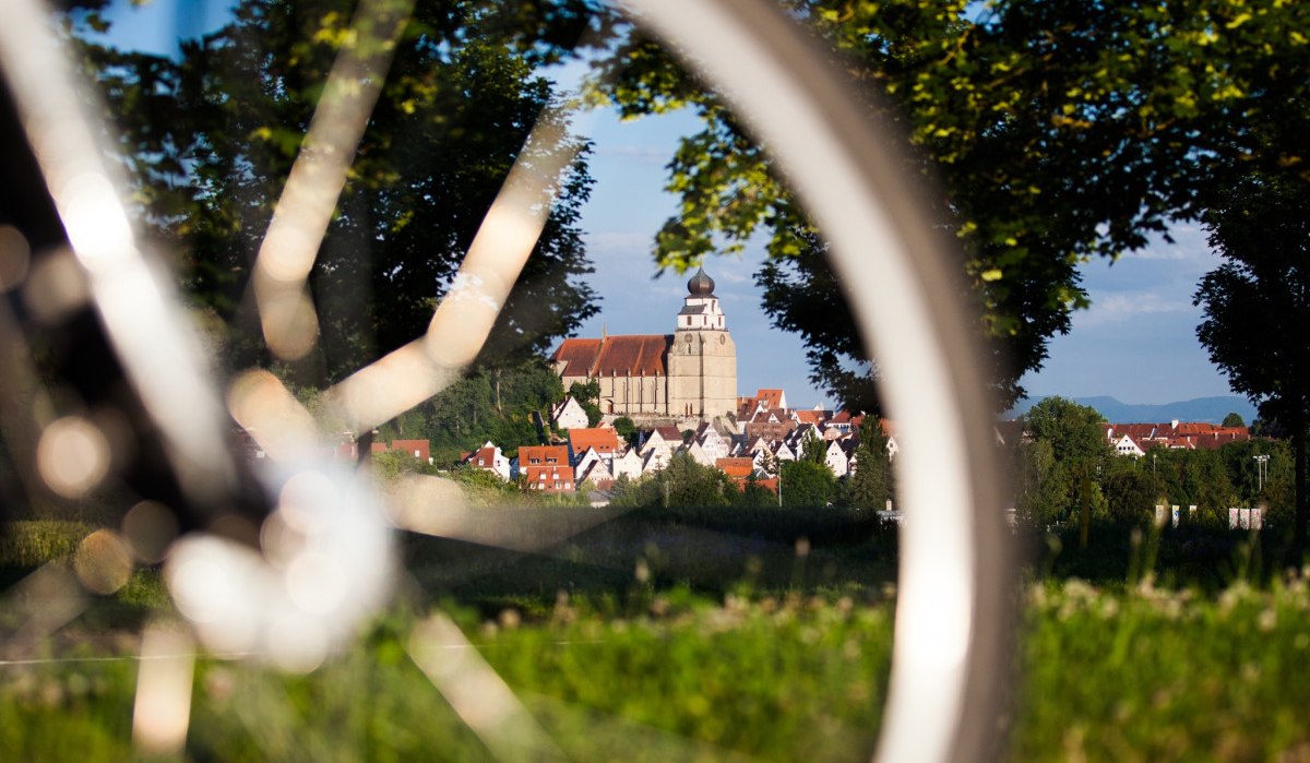 Ein Fahrrad im Vordergrund, durch dessen Speichen eine Kirche in einer Stadt zu sehen ist, umgeben von Bäumen und grüner Landschaft., © Natur.Nah. Schönbuch & Heckengäu Ein Fahrrad im Vordergrund, durch dessen Speichen eine Kirche in einer Stadt zu sehen ist, umgeben von Bäumen und grüner Landschaft., © Natur.Nah. Schönbuch & Heckengäu