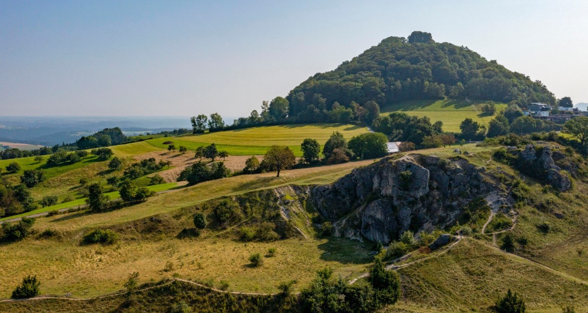 Grüne Hügel und Bäume unter klarem Himmel, mit einem markanten Felsen im Vordergrund. Im Hintergrund ist ein bewaldeter Hügel zu sehen., © TMBW Grüne Hügel und Bäume unter klarem Himmel, mit einem markanten Felsen im Vordergrund. Im Hintergrund ist ein bewaldeter Hügel zu sehen., © TMBW