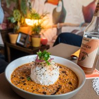 Ein Teller mit Reis und Curry, garniert mit Sesam und Kr&auml;utern, steht auf einem Tisch. Im Hintergrund sind Pflanzen und ein Wandbild mit Tukanen zu sehen., &copy; HerzoginN&uuml;rtingen