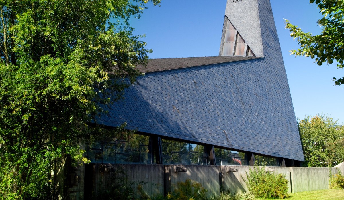 Moderne Kirche mit spitzem Turm und Schieferdach, umgeben von Bäumen und Rasenfläche, unter klarem blauem Himmel., © Waiblingen - Stuttgart-Marketing GmbH Moderne Kirche mit spitzem Turm und Schieferdach, umgeben von Bäumen und Rasenfläche, unter klarem blauem Himmel., © Waiblingen - Stuttgart-Marketing GmbH