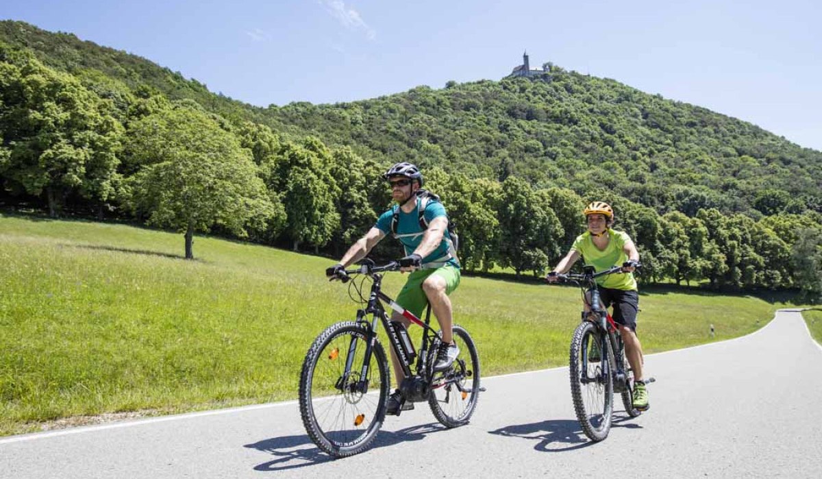 Zwei Mountainbiker fahren auf einer Straße durch eine grüne Landschaft. Im Hintergrund ist ein bewaldeter Hügel mit einem Turm zu sehen., © Schwäbische Alb Tourismusverband e.V. Zwei Mountainbiker fahren auf einer Straße durch eine grüne Landschaft. Im Hintergrund ist ein bewaldeter Hügel mit einem Turm zu sehen., © Schwäbische Alb Tourismusverband e.V.
