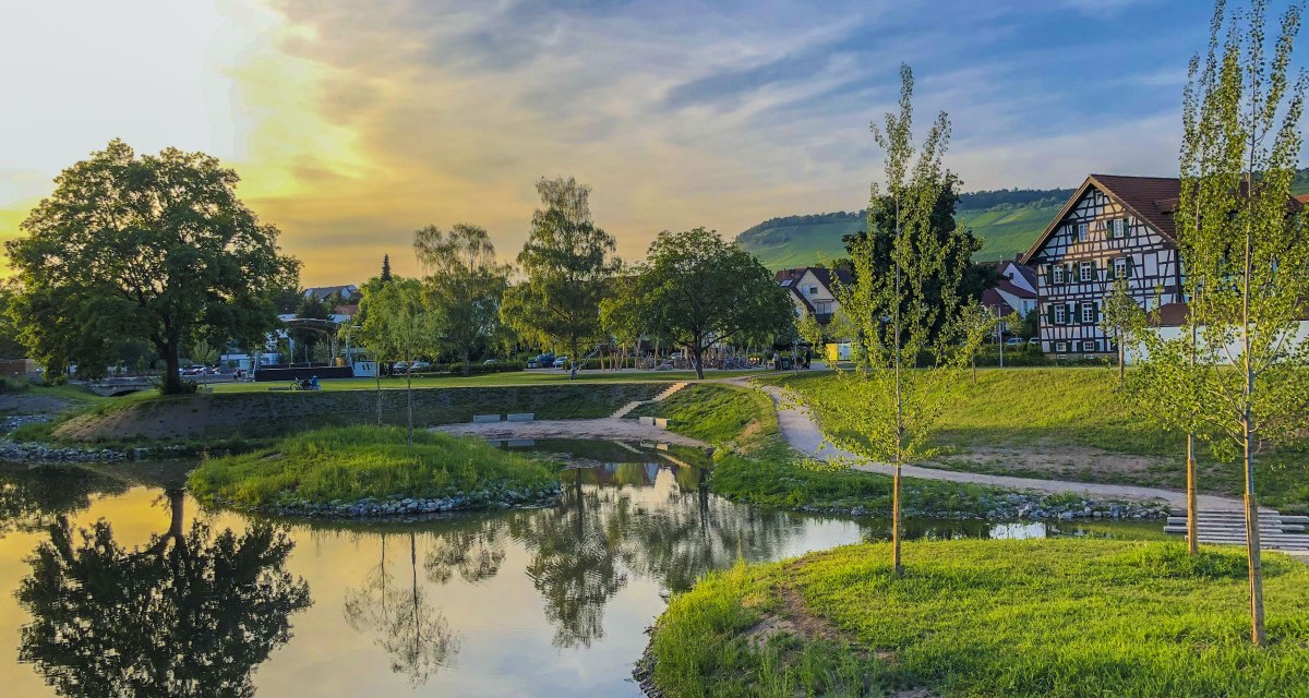 Idyllische Landschaft mit Fluss, Bäumen und Fachwerkhäusern bei Sonnenuntergang in Weinstadt. Der Himmel ist blau mit gelben und orangen Tönen., © Stadt Weinstadt Idyllische Landschaft mit Fluss, Bäumen und Fachwerkhäusern bei Sonnenuntergang in Weinstadt. Der Himmel ist blau mit gelben und orangen Tönen., © Stadt Weinstadt