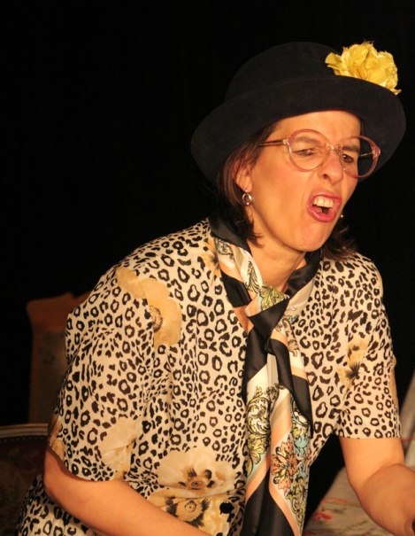Eine Frau mit Brille und Hut mit gelber Blume zieht eine Grimasse. Sie trägt ein Leopardenmuster-Oberteil und einen Schal., © Galli Theater Backnang