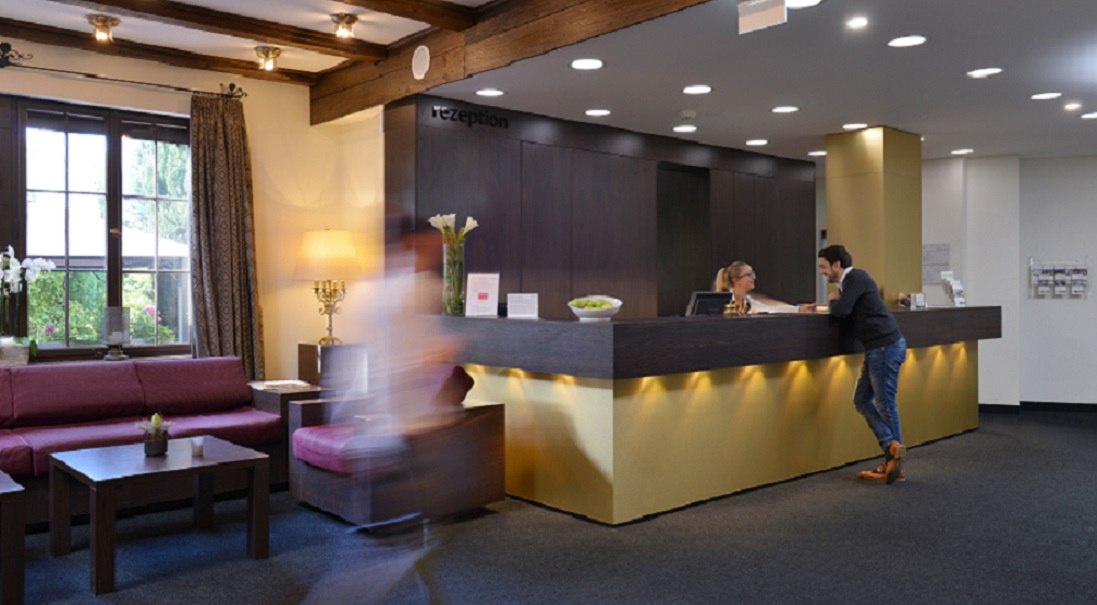 Hotelrezeption mit einem Mann, der mit einer Rezeptionistin spricht. Eine Person geht schnell vorbei. Gemütliche Sitzecke mit Sofa und Tisch., © relexa Waldhotel Schatten Hotelrezeption mit einem Mann, der mit einer Rezeptionistin spricht. Eine Person geht schnell vorbei. Gemütliche Sitzecke mit Sofa und Tisch., © relexa Waldhotel Schatten