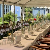 Ein langer Tisch unter einem Pavillon, festlich gedeckt mit Blumenvasen und Geschirr, vor einem blauen Geb&auml;ude im Garten., &copy; cCaf&eacute;Pl&auml;tschwiesle