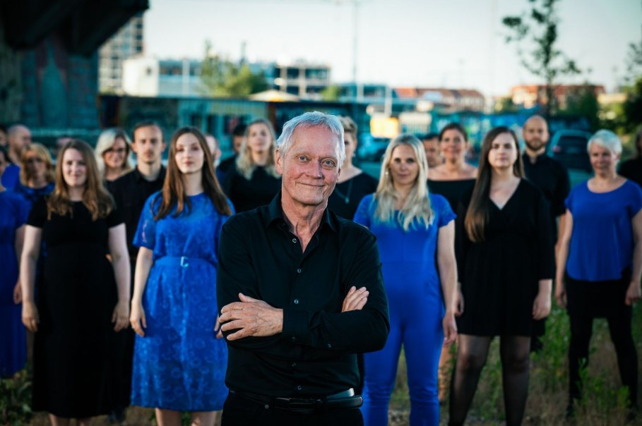 Ein Mann im Vordergrund mit verschränkten Armen, umgeben von einem Chor in blauer und schwarzer Kleidung. Hintergrund zeigt eine urbane Umgebung.
