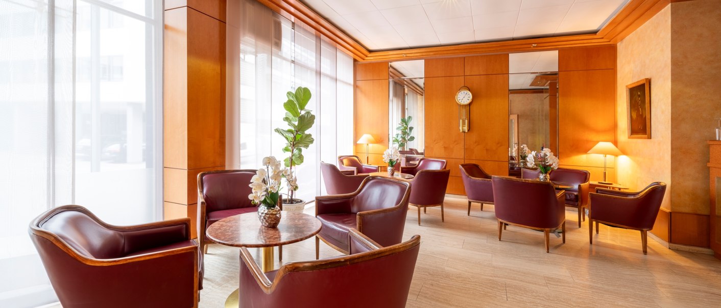 Elegante Lounge mit roten Ledersesseln, Holzwänden und großen Fenstern. Pflanzen und Blumen auf den Tischen, Uhr an der Wand., © PLAZA INN Rieker Stuttgart Hauptbahnhof Elegante Lounge mit roten Ledersesseln, Holzwänden und großen Fenstern. Pflanzen und Blumen auf den Tischen, Uhr an der Wand., © PLAZA INN Rieker Stuttgart Hauptbahnhof