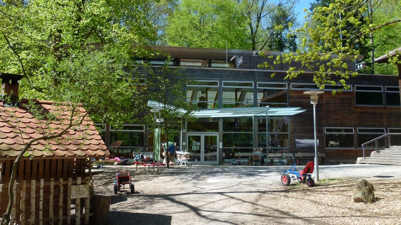Innenhof des Haus des Waldes mit Spielzeugfahrzeugen, Bäumen und einem kleinen Holzhaus. Moderne Glasfassade im Hintergrund., © Haus des Waldes