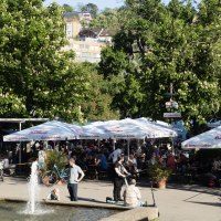 Biergarten im Schlossgarten mit vielen Menschen unter großen Sonnenschirmen. Ein Brunnen im Vordergrund, umgeben von grünen Bäumen und Gebäuden im Hintergrund., © Michele Scognamillo