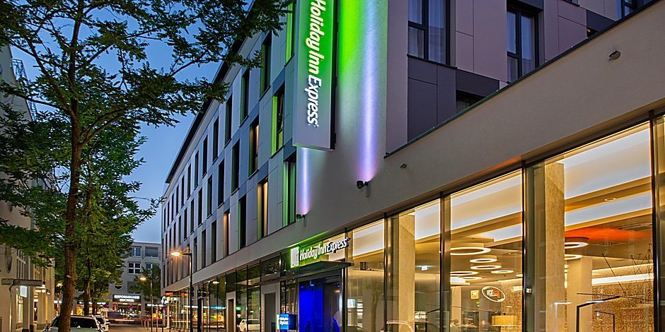 Außenansicht eines modernen Holiday Inn Express Hotels bei Dämmerung, mit beleuchtetem Logo und Glasfassade., © Holiday Inn Express Waiblingen