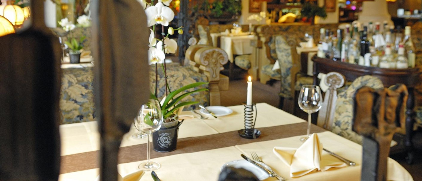 Elegantes Restaurant mit gedecktem Tisch, Orchideen und Kerzenlicht. Im Hintergrund sind weitere Tische und eine Bar zu sehen., © Hotel & Restaurant Beurener Hof Elegantes Restaurant mit gedecktem Tisch, Orchideen und Kerzenlicht. Im Hintergrund sind weitere Tische und eine Bar zu sehen., © Hotel & Restaurant Beurener Hof