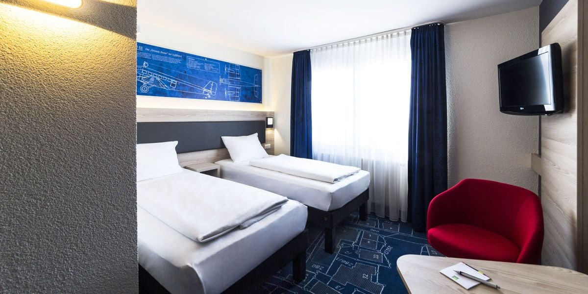 Modernes Hotelzimmer mit zwei Einzelbetten, rotem Sessel, blauem Teppich und Wandkunst. Ein Fenster mit blauen Vorhängen lässt Licht herein., © ibis Styles Filderstadt Messe Stuttgart Modernes Hotelzimmer mit zwei Einzelbetten, rotem Sessel, blauem Teppich und Wandkunst. Ein Fenster mit blauen Vorhängen lässt Licht herein., © ibis Styles Filderstadt Messe Stuttgart