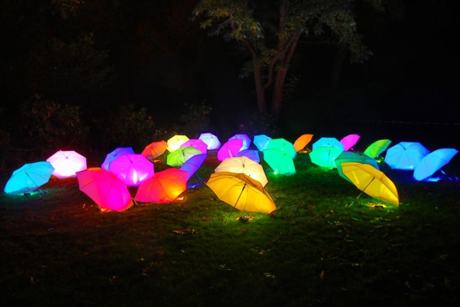 Leuchtende bunte Regenschirme liegen bei Nacht auf einer Wiese, umgeben von B&auml;umen. Die Schirme strahlen in verschiedenen Farben., &copy; copyright: Marc Sansone