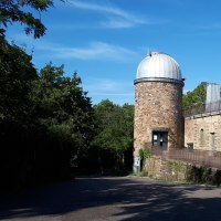 Eine Sternwarte mit runder Kuppel und Eingang, umgeben von Bäumen und blauem Himmel., © SMG Eine Sternwarte mit runder Kuppel und Eingang, umgeben von Bäumen und blauem Himmel., © SMG