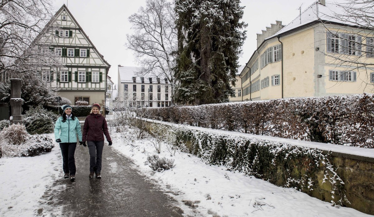 Zwei Personen spazieren auf einem verschneiten Weg in einer winterlichen Stadt. Im Hintergrund sind ein Fachwerkhaus und schneebedeckte Bäume zu sehen., © Schwäbische Alb Tourismusverband e.V.