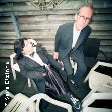 Zwei Personen in formeller Kleidung sitzen auf wei&szlig;en Plastikst&uuml;hlen vor einer Holzwand. Eine Person sitzt entspannt, die andere lehnt sich nach vorne., &copy; links im Bild