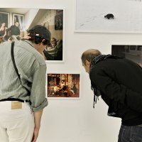 Zwei Personen betrachten aufmerksam Fotos an einer weißen Galeriewand. Die Bilder zeigen verschiedene Szenen, darunter Menschen und eine Schneelandschaft., © Palermo Galerie