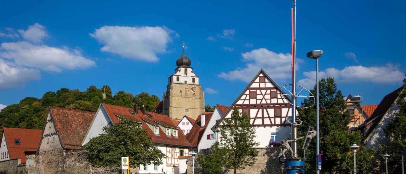 Die Stiftskirche Herrenberg erhebt sich über Fachwerkhäuser, umgeben von Bäumen und einem klaren blauen Himmel mit wenigen Wolken., © SMG Achim Mende Die Stiftskirche Herrenberg erhebt sich über Fachwerkhäuser, umgeben von Bäumen und einem klaren blauen Himmel mit wenigen Wolken., © SMG Achim Mende