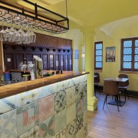 Ein gem&uuml;tliches Restaurant mit gelben W&auml;nden, einer Bar mit bunten Fliesen und h&auml;ngenden Weingl&auml;sern. Im Hintergrund sind Tische und St&uuml;hle zu sehen.