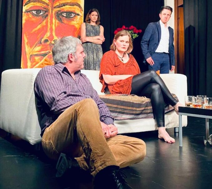 Theaterszene mit vier Personen in einem Wohnzimmer. Eine Person sitzt auf dem Boden, zwei auf dem Sofa, eine steht im Hintergrund. Ein großes Gesichtsgemälde ist sichtbar., © Stadt Sindelfingen