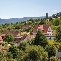 Ein malerisches Dorf mit traditionellen Fachwerkhäusern, umgeben von grünen Hügeln und Bäumen, unter einem klaren blauen Himmel., © SMG, Achim Mende Ein malerisches Dorf mit traditionellen Fachwerkhäusern, umgeben von grünen Hügeln und Bäumen, unter einem klaren blauen Himmel., © SMG, Achim Mende