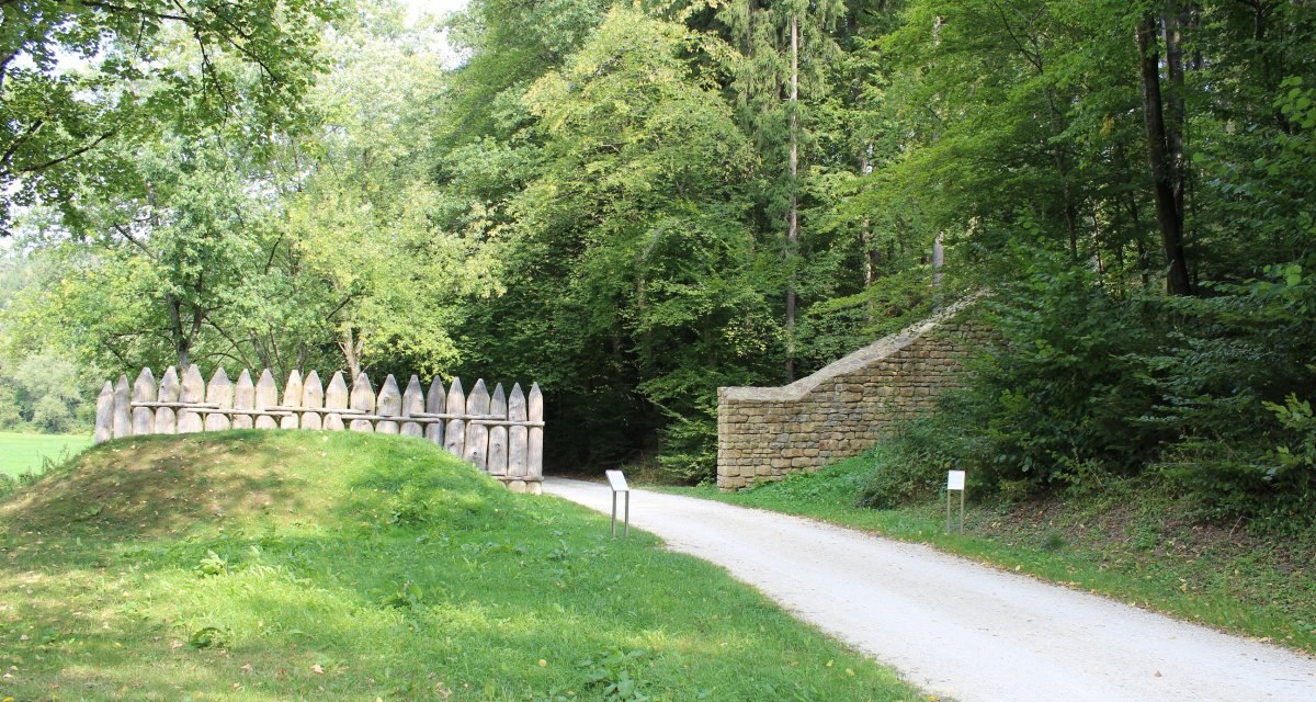 Rekonstruktion eines römischen Limesabschnitts im Wald mit Palisaden und Mauer, umgeben von Bäumen und einem Weg., © Foto Harald Messerschmidt Rekonstruktion eines römischen Limesabschnitts im Wald mit Palisaden und Mauer, umgeben von Bäumen und einem Weg., © Foto Harald Messerschmidt