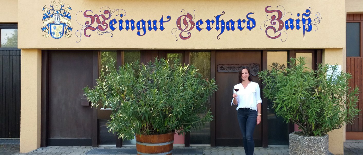 Eine Frau steht vor dem Weingut Gerhard Zaiß, hält ein Glas Rotwein und lächelt. Das Gebäude hat eine dekorative Beschriftung und Pflanzen im Vordergrund., © Simone Mack Eine Frau steht vor dem Weingut Gerhard Zaiß, hält ein Glas Rotwein und lächelt. Das Gebäude hat eine dekorative Beschriftung und Pflanzen im Vordergrund., © Simone Mack