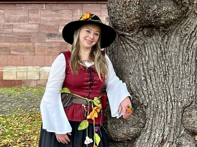 Eine Frau in mittelalterlicher Kleidung mit Blumen am Gürtel lehnt lächelnd an einem großen Baum vor einer Steinmauer., © Stadtverwaltung Weil der Stadt Eine Frau in mittelalterlicher Kleidung mit Blumen am Gürtel lehnt lächelnd an einem großen Baum vor einer Steinmauer., © Stadtverwaltung Weil der Stadt