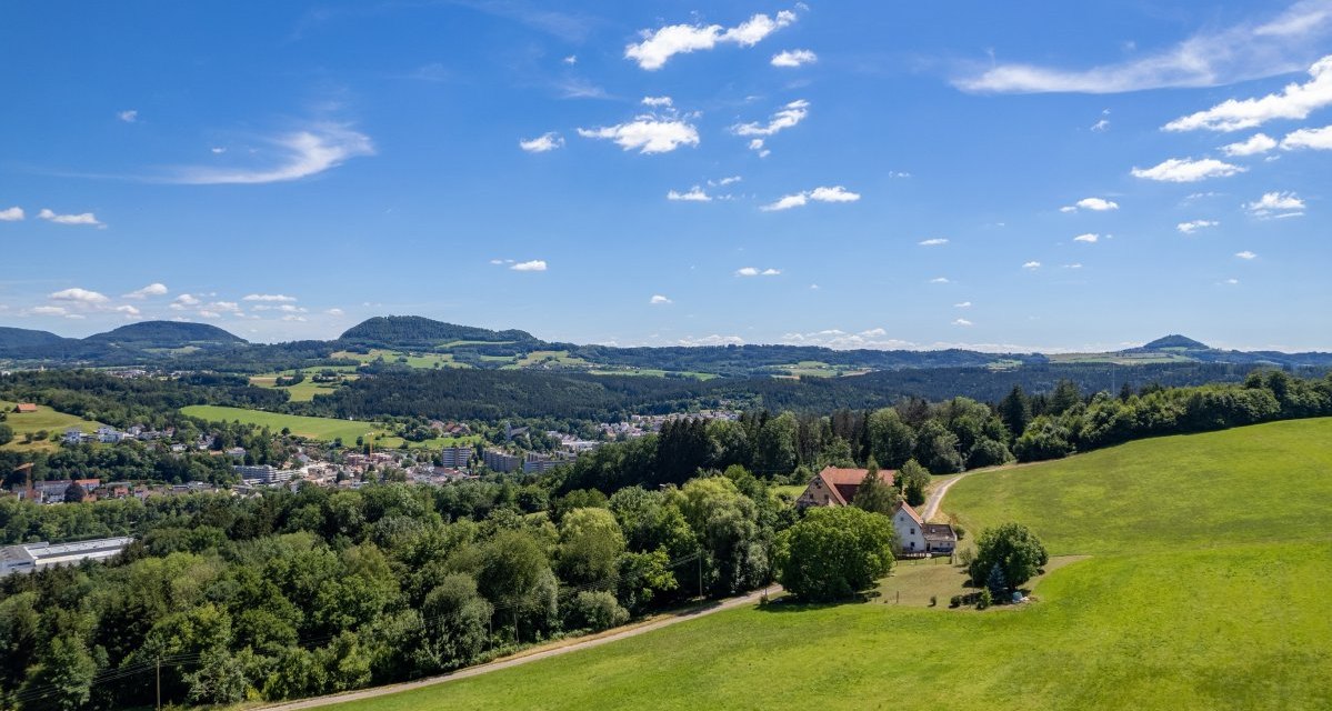 Weite Landschaft mit grünen Hügeln, Wiesen und einem Bauernhof. Im Hintergrund sind bewaldete Hügel und ein klarer blauer Himmel zu sehen., © Foto Thomas Zehnder Weite Landschaft mit grünen Hügeln, Wiesen und einem Bauernhof. Im Hintergrund sind bewaldete Hügel und ein klarer blauer Himmel zu sehen., © Foto Thomas Zehnder