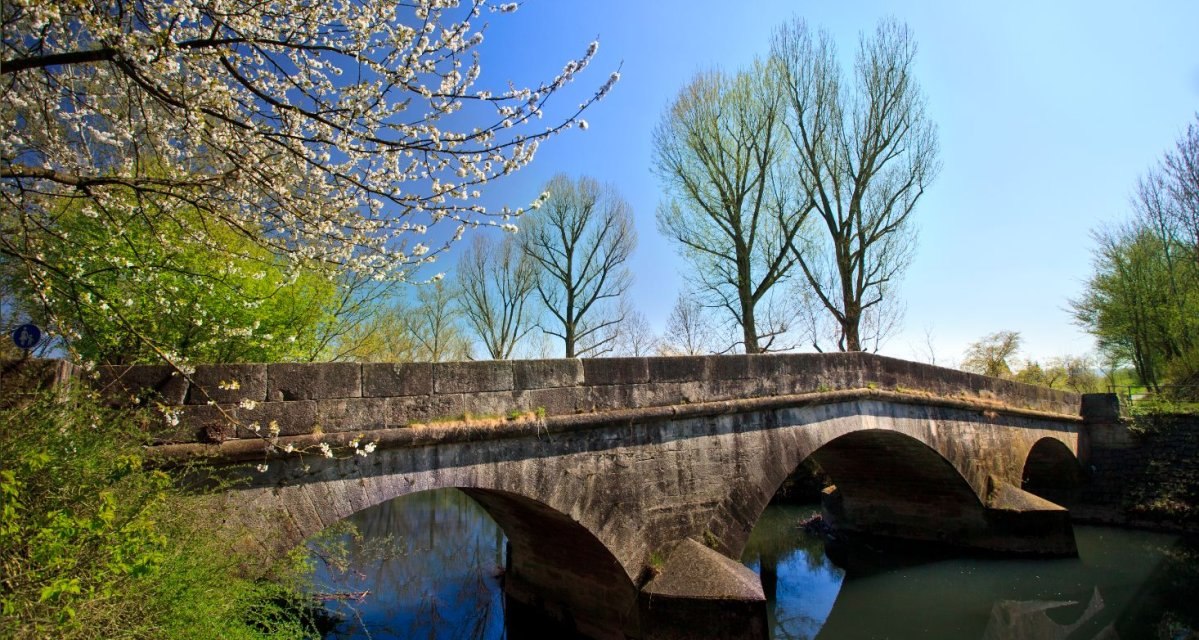 Steinbrücke mit zwei Bögen über einen Fluss, umgeben von blühenden Bäumen und blauem Himmel., © Waiblingen - Stuttgart-Marketing GmbH Steinbrücke mit zwei Bögen über einen Fluss, umgeben von blühenden Bäumen und blauem Himmel., © Waiblingen - Stuttgart-Marketing GmbH
