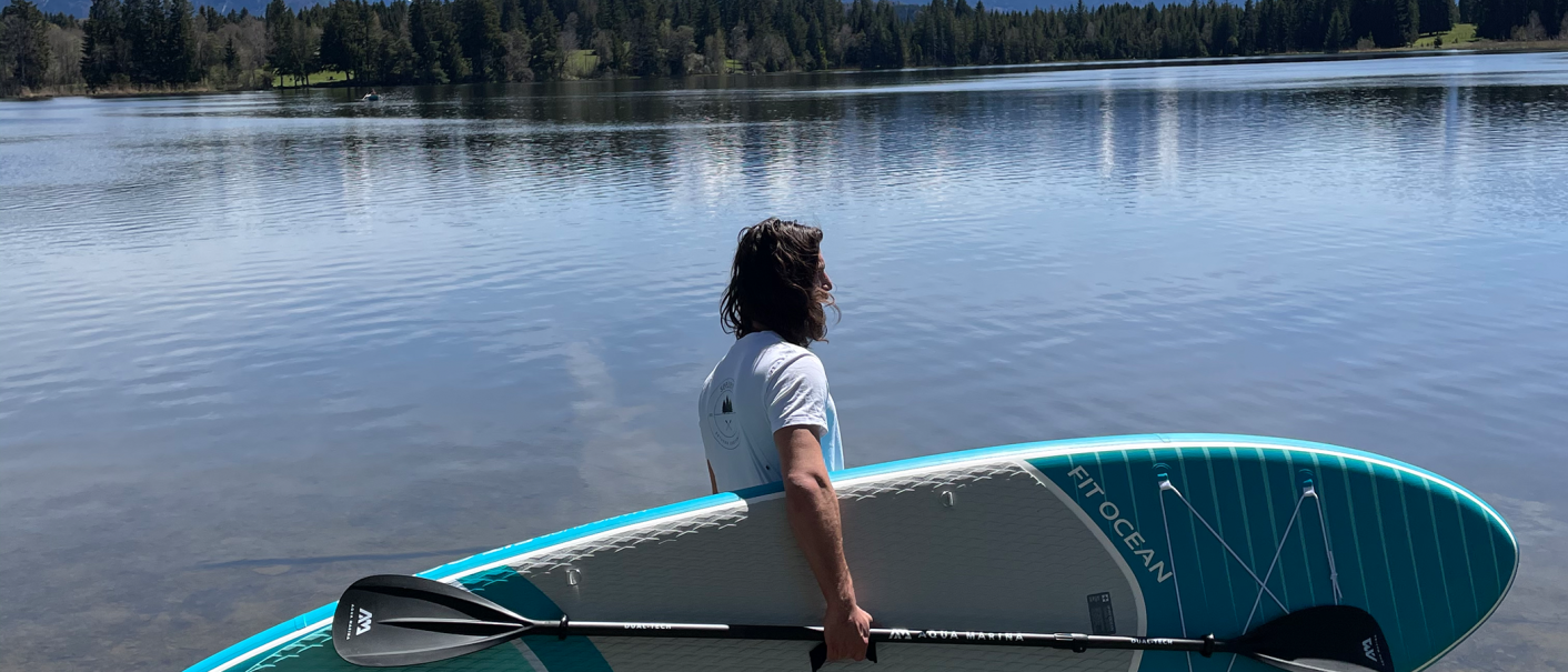 Eine Person steht am Ufer eines Sees mit einem SUP-Board und Paddel, umgeben von Wald und ruhigem Wasser., © seezeit GmbH Eine Person steht am Ufer eines Sees mit einem SUP-Board und Paddel, umgeben von Wald und ruhigem Wasser., © seezeit GmbH