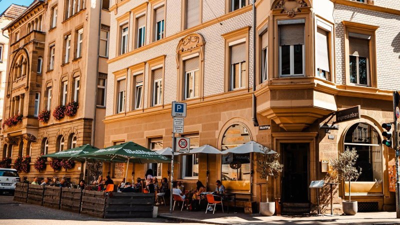 Stra&szlig;encaf&eacute; mit Sonnenschirmen vor einem historischen Geb&auml;ude. Menschen sitzen drau&szlig;en, genie&szlig;en das Wetter. Verkehrsschilder und eine Vespa sind sichtbar., &copy; Stuttgart Marketing GmbH, Sarah Schmid