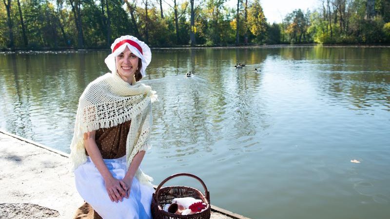 Eine Frau in historischem Kost&uuml;m sitzt l&auml;chelnd am Ufer eines Sees. Sie tr&auml;gt eine Haube und einen Umhang, neben ihr steht ein Korb., &copy; &copy; T&E, Reiner Pfisterer