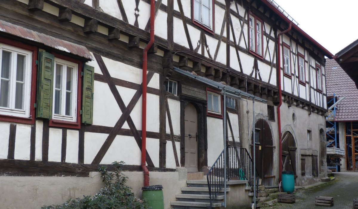 Historisches Fachwerkhaus mit Holzbalken, roten Fensterläden und grünen Regentonnen. Treppe führt zur Eingangstür. Hof mit Pflastersteinen., © Land der 1000 Hügel - Kraichgau-Stromberg