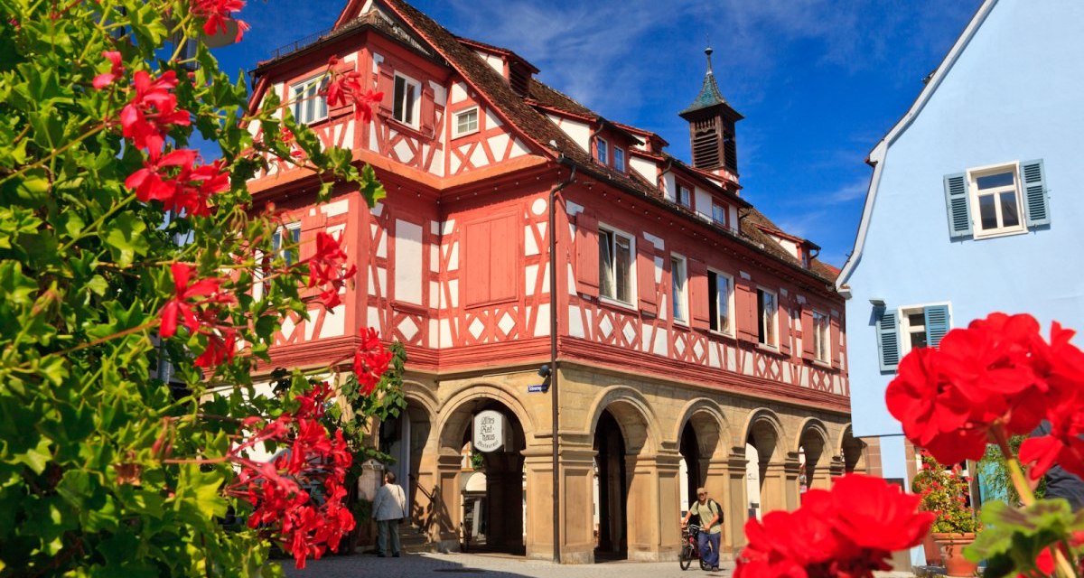 Ein historisches Fachwerkhaus mit roter Fassade und Arkaden, umgeben von roten Blumen, unter einem strahlend blauen Himmel., © WTM GmbH Waiblingen Ein historisches Fachwerkhaus mit roter Fassade und Arkaden, umgeben von roten Blumen, unter einem strahlend blauen Himmel., © WTM GmbH Waiblingen