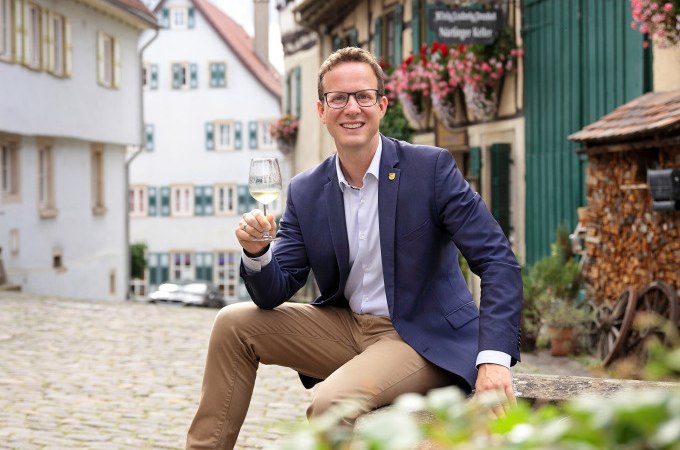 Ein Mann in einem blauen Sakko sitzt in einer malerischen Altstadt und hält ein Glas Wein. Im Hintergrund sind Fachwerkhäuser und Blumen zu sehen., © Stadt Nürtingen Ein Mann in einem blauen Sakko sitzt in einer malerischen Altstadt und hält ein Glas Wein. Im Hintergrund sind Fachwerkhäuser und Blumen zu sehen., © Stadt Nürtingen