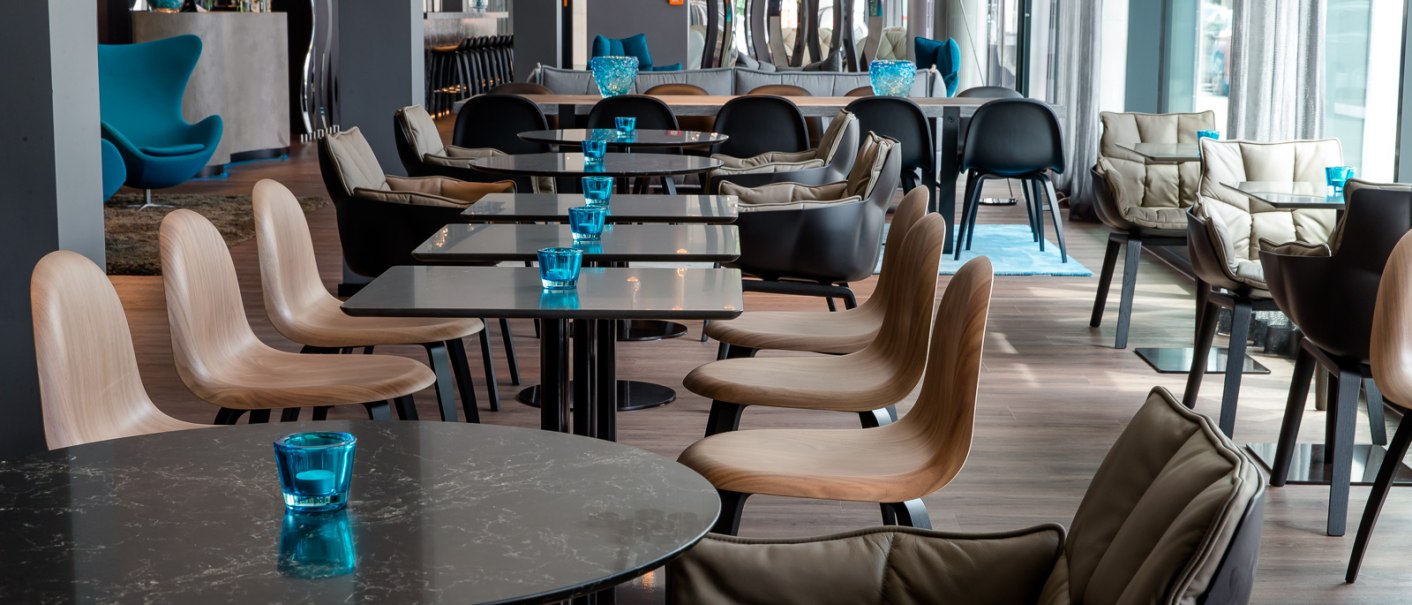 Moderner Frühstücksbereich mit eleganten Tischen, Stühlen und blauen Akzenten. Stilvolle Beleuchtung und große Fenster sorgen für eine einladende Atmosphäre., © Motel One Stuttgart-Bad Cannstatt