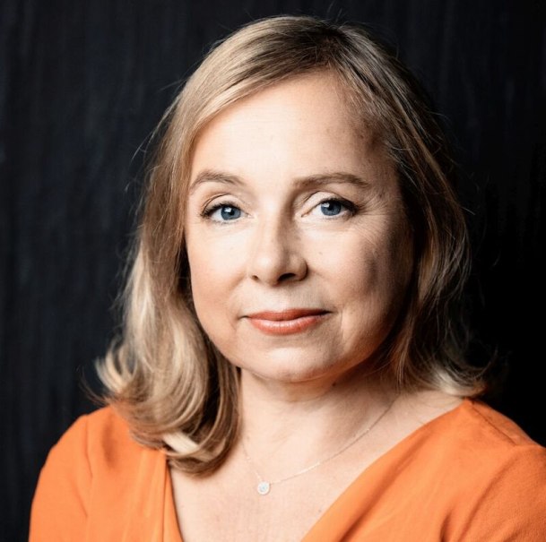 Frau mit blonden Haaren und orangefarbener Kleidung blickt in die Kamera, vor dunklem Hintergrund., © Stadthalle Leonberg Frau mit blonden Haaren und orangefarbener Kleidung blickt in die Kamera, vor dunklem Hintergrund., © Stadthalle Leonberg