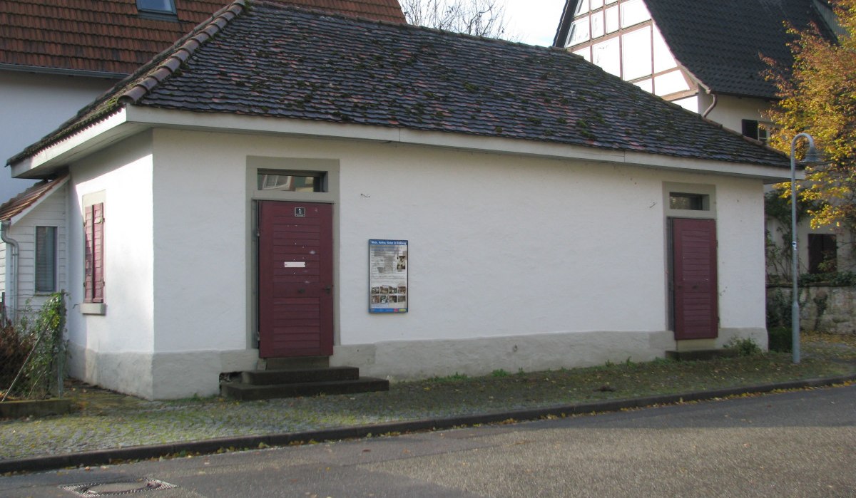 Kleines, weißes Gebäude mit roten Türen und Ziegeldach, umgeben von Wohnhäusern. Ein Schild ist an der Wand angebracht., © Land der 1000 Hügel - Kraichgau-Stromberg