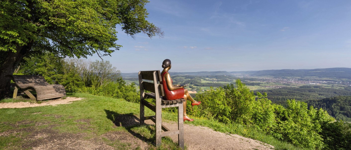 Eine Skulptur einer Frau sitzt auf einer übergroßen Bank und blickt auf eine weite, grüne Landschaft unter blauem Himmel., © Stuttgart-Marketing GmbH, Martina Denker Eine Skulptur einer Frau sitzt auf einer übergroßen Bank und blickt auf eine weite, grüne Landschaft unter blauem Himmel., © Stuttgart-Marketing GmbH, Martina Denker
