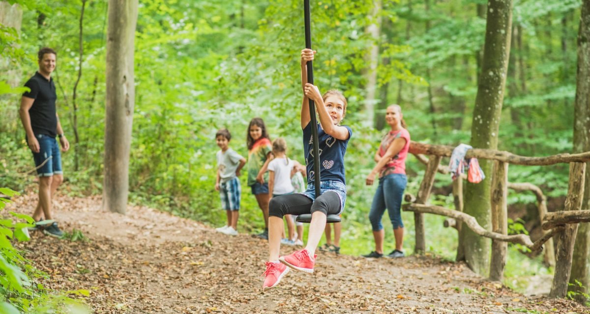 Ein Kind schwingt an einer Seilrutsche im Wald, umgeben von anderen Kindern und Erwachsenen, die zuschauen. Der Spielplatz liegt im Grünen. Ein Kind schwingt an einer Seilrutsche im Wald, umgeben von anderen Kindern und Erwachsenen, die zuschauen. Der Spielplatz liegt im Grünen.