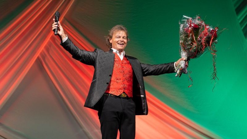 Ein Mann in einem schwarzen Anzug mit roter Weste steht auf einer B&uuml;hne, h&auml;lt ein Mikrofon und einen Blumenstrau&szlig;. Der Hintergrund ist in Rot und Gr&uuml;n beleuchtet., &copy; Burkauer Veranstaltungsservice