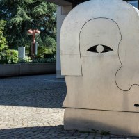Eine große Metallskulptur in Form eines stilisierten Kopfes steht auf einem gepflasterten Platz, umgeben von Grünpflanzen., © Fotoknobi