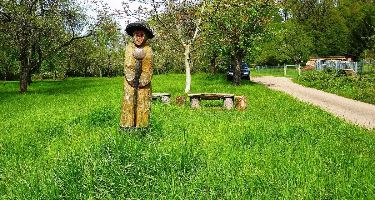 Holzfigur eines Pilgers auf einer grünen Wiese, umgeben von Bäumen. Ein Weg und eine Bank sind im Hintergrund zu sehen., © Plochingen Holzfigur eines Pilgers auf einer grünen Wiese, umgeben von Bäumen. Ein Weg und eine Bank sind im Hintergrund zu sehen., © Plochingen