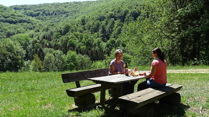 Zwei Personen sitzen an einem Holztisch auf einer grünen Wiese, umgeben von Wald. Sie genießen ein Picknick bei sonnigem Wetter., © Kurverwaltung Beuren, Kaplan