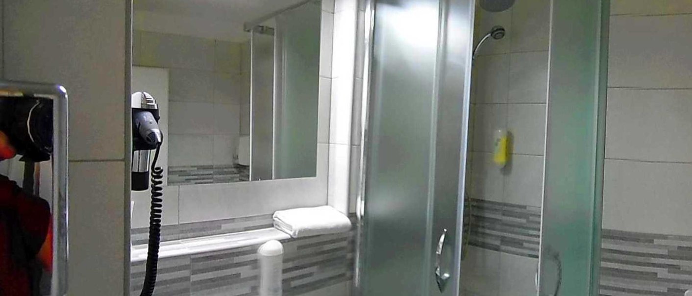 Modernes Badezimmer mit Dusche, Spiegel, Haartrockner und Handtuch. Die Wände sind mit grauen Fliesen dekoriert., © Hotel Arcis Modernes Badezimmer mit Dusche, Spiegel, Haartrockner und Handtuch. Die Wände sind mit grauen Fliesen dekoriert., © Hotel Arcis